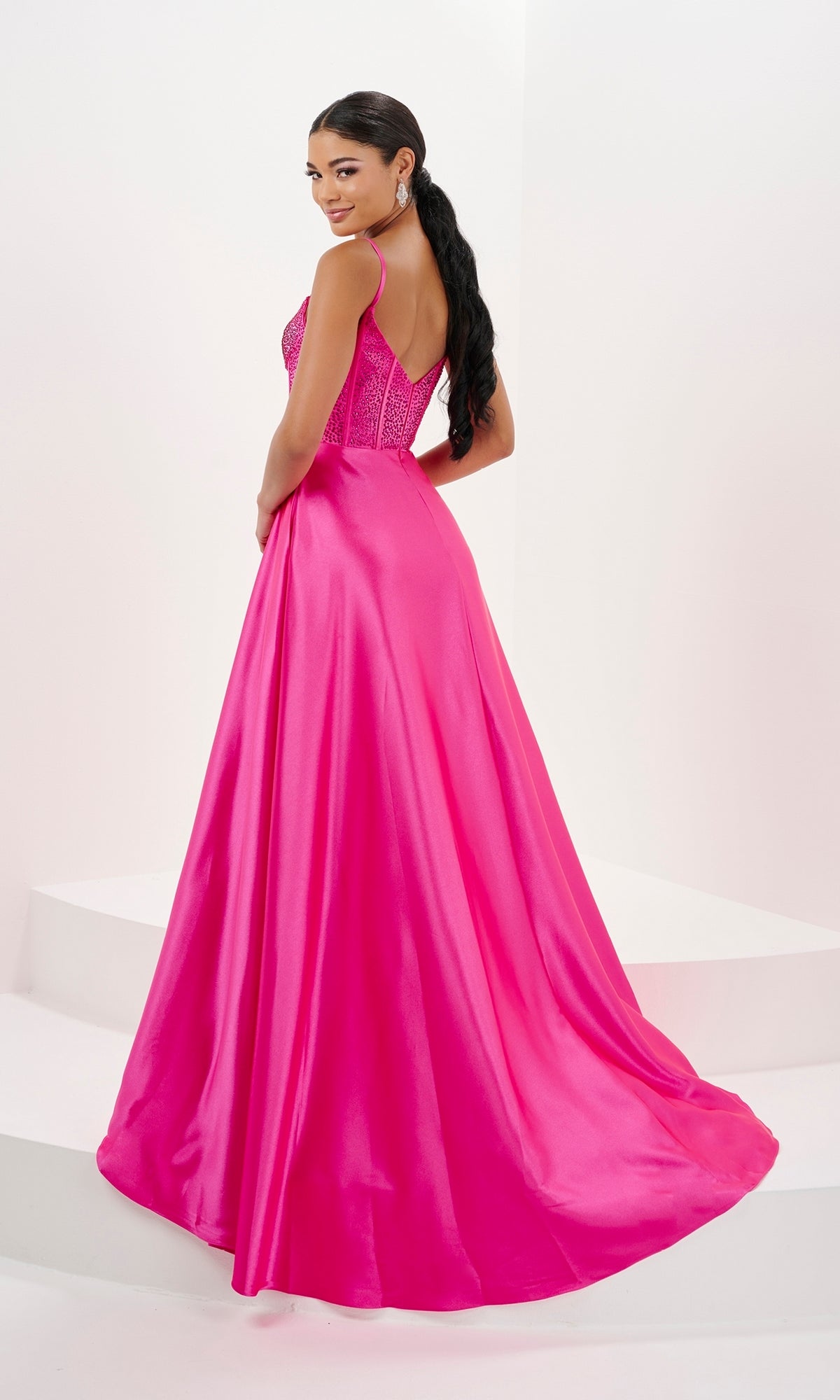 A woman stands on a white platform wearing the Tiffany Long Jersey A-Line Corset Prom Dress 16101 in bright pink, featuring a floor-length skirt and corset bodice, looking over her shoulder.
